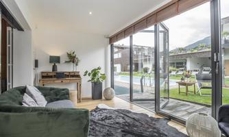 A modern living room with a green sofa and a desk. Large windows offer a view of the garden and the swimming pool.