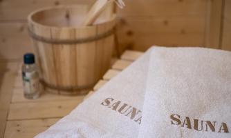 A quiet sauna area with two white towels that have "SAUNA" printed on them. In the background, there is a wooden bucket and a bottle of essential oils.