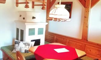 A cozy dining area with a round table and wooden furniture. The table is decorated with a red cloth and there is a traditional stove in the corner.