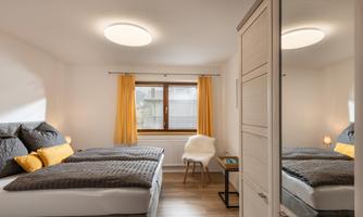 A modern bedroom with two beds, elegant pillows, and a large mirror. The windows are adorned with yellow curtains, and the room is bright and inviting.