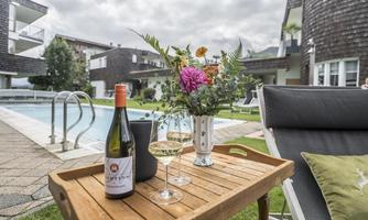 A table with a bottle of wine and glasses stands next to a pool. In the background, modern buildings and colorful flowers can be seen.