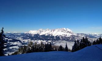 A stunning view of snow-covered mountains under a clear blue sky. The foreground features a snowy landscape with coniferous trees.
