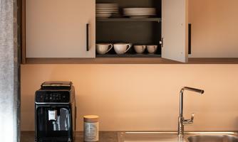 A modern kitchen with a black coffee machine and a shiny sink. In the upper cabinet, dishes and glasses are neatly arranged.