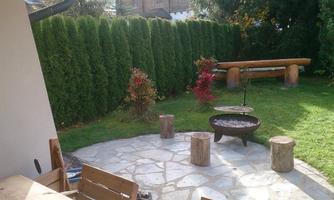 A cozy garden with a stone floor and a grill area. Surrounded by green hedges and colorful plants.