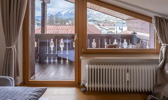 A cozy room with large windows that look out onto a balcony and the mountains. The wooden floor and the curtains create a warm atmosphere.
