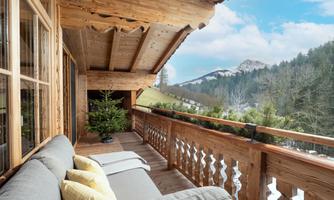 A cozy wooden porch with a comfortable couch and cushions. In the background, green mountains and a blue sky can be seen.