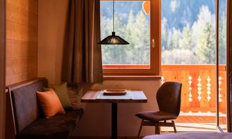 A cozy dining area in a wooden house with a table and a bench. Through the window, a picturesque landscape with trees is visible.