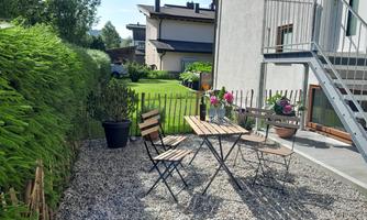 A cozy outdoor area with a wooden table and chairs. Surrounded by green foliage and a small garden.