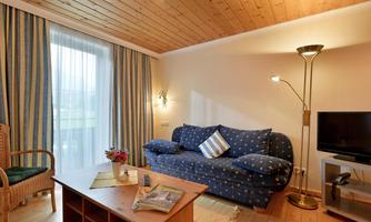 A cozy living room with a blue sofa and a floor lamp. Large windows let in plenty of light, and there is a wooden shelf with a small flower vase.