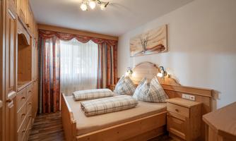 A cozy bedroom with wooden furniture and two beds. The windows are well-lit with curtains and a modern wall mural.
