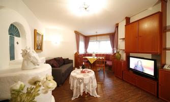 A cozy living room with a gray sofa and a round table. The room features a television and a small kitchen with wooden cabinets.