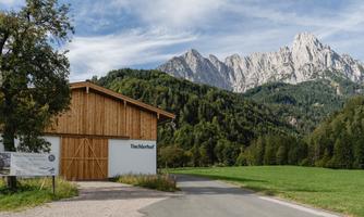 A modern wooden building stands along a path in a green landscape. In the background, majestic mountains rise under a blue sky.