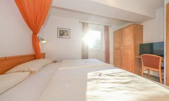 A bright bedroom with two single beds and warm color details. A window lets sunlight in, and there is a desk as well as a television in the room.