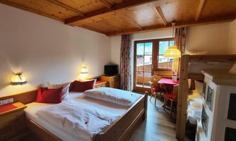 A cozy room with a double bed and wooden details. It has large windows and a small dining area.