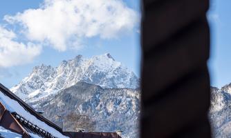 A snow-covered mountain landscape with clear blue sky and clouds. In the foreground, part of a roof is visible.