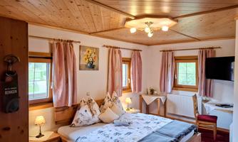 A cozy bedroom with a large bed and wooden ceiling. The walls are pleasantly decorated and there is a desk with a chair.