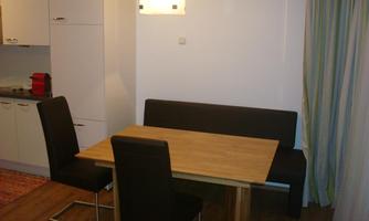 A simple dining room with a wooden table and black chairs. The walls are in light colors, and there is a window with curtains.