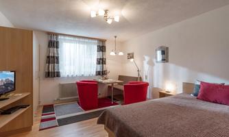 A cozy room with a double bed, a table, and red chairs. Large windows provide plenty of daylight.