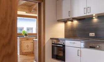 A modern kitchen with white cabinets and an oven. In the background, another room with wooden furniture and plants is visible.