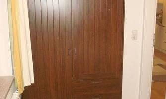 A simple wooden cabinet stands against the wall of a room. Next to it is a bed with a yellow-white bedspread.