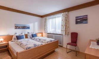 A cozy bedroom with two single beds and bright colors. A chair and a desk complement the room, which has plenty of daylight.