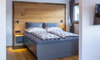 A modern bedroom with a large double bed and a wooden back wall. The room has light walls and a warm wooden floor, with windows that let in light.