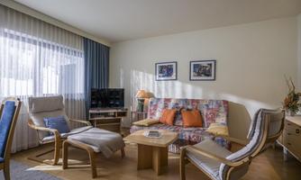 A cozy living room with a colorful sofa and comfortable armchairs. Large windows with curtains provide plenty of natural light.