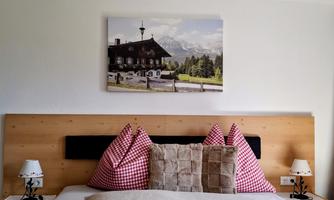 A cozy bedroom with a large bed and colorful pillows. On the wall hangs a picture of a mountain cabin in nature.