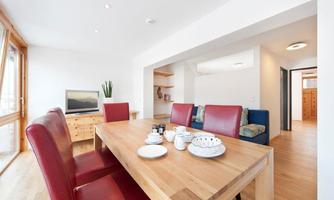 A modern living-dining room with a wooden table and red chairs. In the background, a TV and a cozy seating area can be seen.