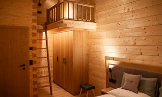 A cozy wooden house room with a large bed and a wardrobe. A staircase leads to a small gallery above.