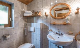 A modern bathroom with white tiles and wood details. It features a sink, a toilet, and a round mirror.