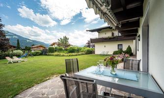 A cozy outdoor area with a table and chairs. In the background, a beautiful garden stretches out with views of the mountains and the blue sky.