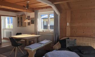 A cozy living room with wooden walls and a large window. There is a dining table, chairs, and a comfortable couch.