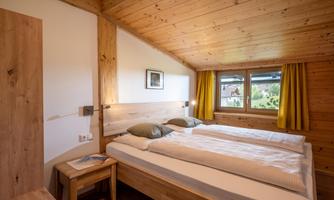 A cozy bedroom with wooden paneling and two single beds. Large windows with yellow curtains provide a view outside.