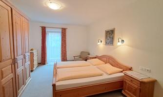 A bright bedroom with two single beds and wooden wardrobes. The room has large windows and a cozy atmosphere.