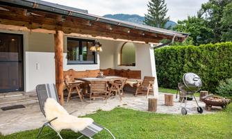 A cozy terrace with wooden furniture and a grill. In the background, trees and the house with a green lawn can be seen.