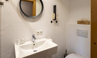 A modern bathroom with a sink, a round mirror, and a toilet. The walls are in neutral colors and the room feels bright and friendly.