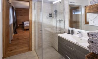 A modern bathroom with a glass shower and an elegant washbasin. In the background, a cozy bedroom can be seen.
