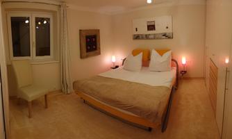 A cozy bedroom with a double bed and two pillows. The walls are in light colors and there is a chair as well as soft lighting.