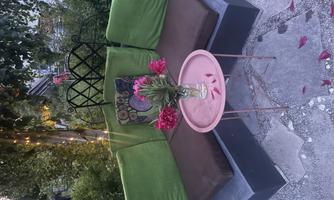A cozy outdoor seating area with green cushions and a small pink table. A vase with blooming flowers sits on the table, surrounded by a green garden landscape.