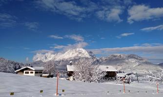 A winter landscape with snow-covered mountains and a clear blue sky. In the foreground, there are some charming houses in the snow.