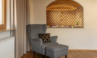 A cozy armchair with an ottoman is placed in a bright room. In the background, there is an architectural niche with a decorative grille visible.
