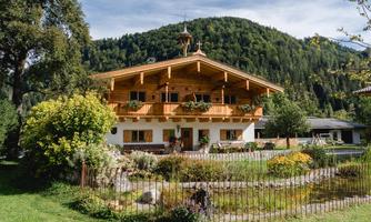 A traditional farmhouse with a beautiful garden and a view of the surrounding mountains. The balconies are adorned with flowers, and the scene radiates tranquility and a connection to nature.