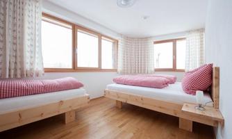 A bright room with two single beds and red checkered blankets. Large windows provide plenty of daylight and a cozy atmosphere.