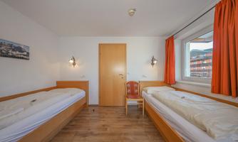 A simple room with two single beds and a chair. The walls are light and the windows have orange curtains.