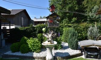 A beautiful garden with a fountain and well-kept greenery. In the background, wooden houses and a birdhouse can be seen.