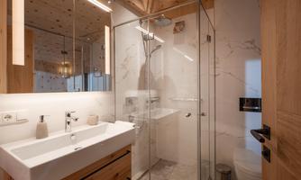 A modern bathroom with a glass shower cubicle and a white vanity unit. The walls are light-tiled and there are wooden elements.