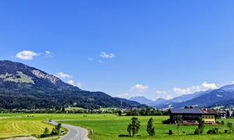 An idyllic landscape with green meadows and gentle hills. In the background, majestic mountains and a clear blue sky are visible.