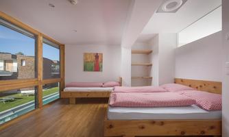 A modern bedroom with two single beds and wooden furniture. Large windows provide plenty of natural light and a view of the outdoors.