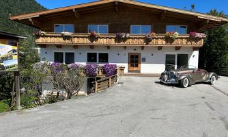 A beautiful chalet with flower-filled balconies and a modern car in front. Surrounded by green mountains and clear skies.